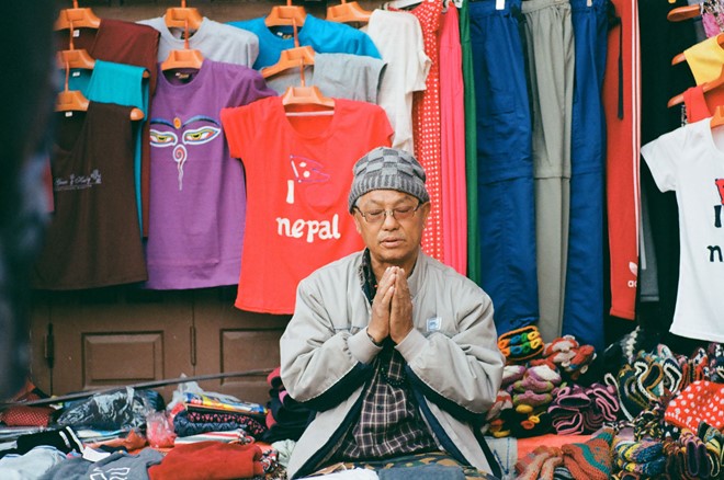 Description: Một người buôn bán quần áo ở Kathmandu hướng về bảo tháp để cầu nguyện. Với người dân ở đây, việc thực hành hàng ngày tự nhiên như hơi thở.